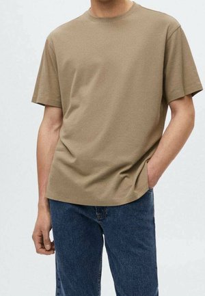 Mann trägt ein schlichtes beige T-Shirt mit kurzen Ärmeln und blaue Jeans, eine Hand in der Tasche, vor hellem Hintergrund.