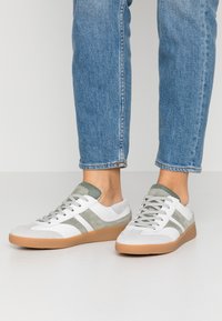 Person wearing white and gray sneakers with gum soles and blue jeans standing on a white floor.