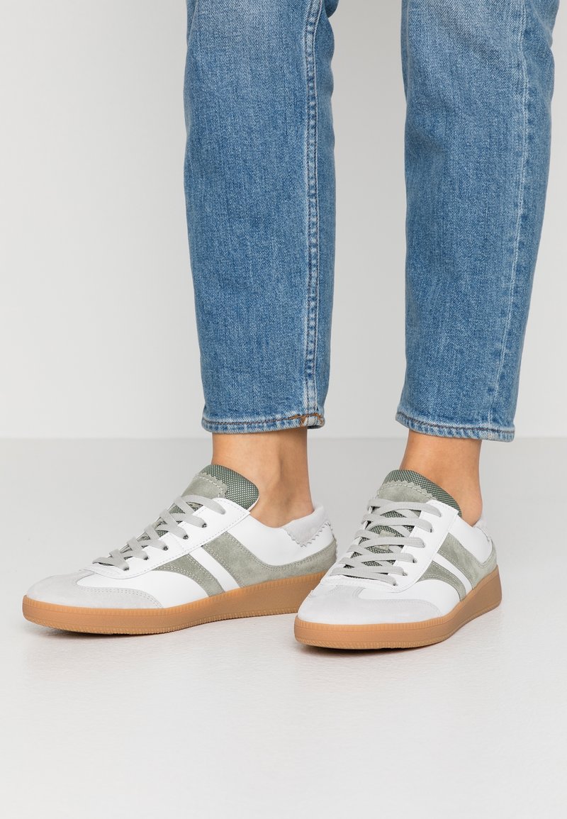 Person wearing white and gray sneakers with gum soles and blue jeans standing on a white floor.