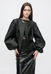 Jachetă cropped din satin negru, cu mâneci voluminoase, având o dantelă la decolteu, purtată alături de o fustă din piele mulată.