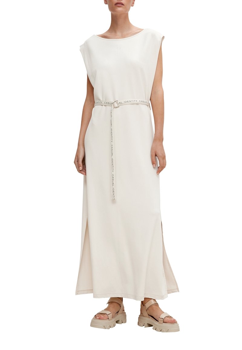 Vestido largo de color crema con mangas anchas, cintura ceñida con un cinturón que presenta texto de logo y aberturas laterales. Combinado con sandalias gruesas de color marrón claro.