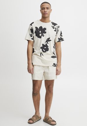 Light beige cotton t-shirt with large black floral pattern, paired with cream shorts and brown slide sandals. Simple, casual design.