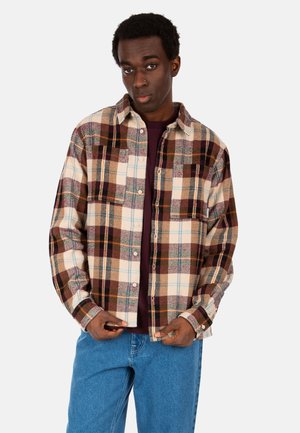 Junger Mann mit dunkler Haut und kurzen lockigen Haaren trägt ein braun-beiges kariertes Hemd über einem dunklen Hemd und blauen Jeans und richtet den Saum seines Hemdes.