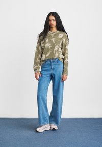 Alyvų žalia megztinis su gėlių raštu, suglamžyta tekstūra; derinamas su šviesiai mėlynomis plačiomis kelnėmis. Modelis vilki šviesias sportines batus.