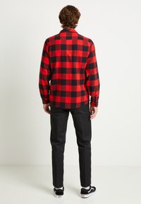 Camisa de franela de cuadros rojos y negros con mangas largas, vista trasera. Combinada con pantalones negros y zapatillas negras. Tejido suave y texturizado, puños con botones.