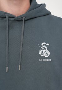 Hettegenser i mørkegrå bomull. Har en hvit slange-logo og teksten "HAN KJØBENHAVN" på fronten. Snorene har sølvfargede spisser.