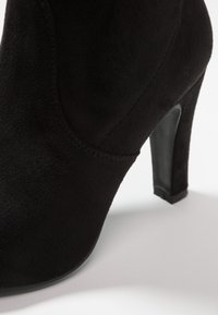 Botte à talon haut en daim noir avec une texture lisse, un bout effilé et un talon poli. Détails de couture le long de la couture.