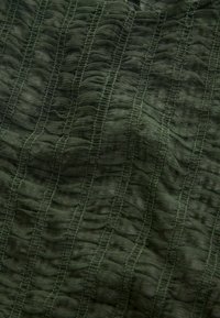 Teksturowana oliwkowozielona tkanina z pionowymi plisami i drobnym przeszyciem, tworząca wzorzystą powierzchnię z głębią i zróżnicowaniem.