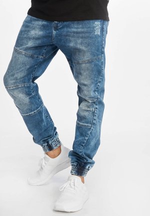 Persona che indossa jeans jogger di denim blu con polsini elastici e scarpe da ginnastica bianche, in piedi su sfondo bianco.