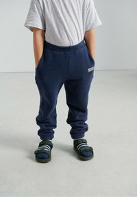 Tumši zili flīsa sporta bikses ar elastīgu jostasvietu un sānu kabatām. Kombinētas ar zaļām sandālēm un tumši ziliem zeķēm ar baltām svītrām.