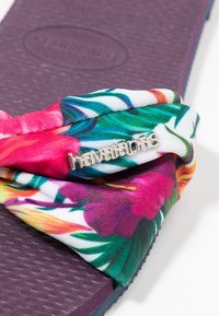 Flip-flop med en texturerad lila gummisula och ett livligt blommönstrat tygband med en silverfärgad "havaianas"-logotyp.