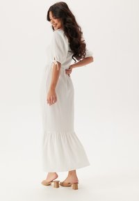 Witte maxi-jurk van gestructureerde stof, met puffmouwen en een uitlopende zoom. Gecombineerd met beige blokhak sandalen.