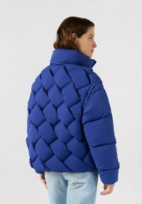 Blauer Puffermantel mit hohem Kragen, der ein gestepptes Diamantmuster, eine weiche Textur und eine entspannte Passform aufweist. Kombiniert mit hellen Jeans.