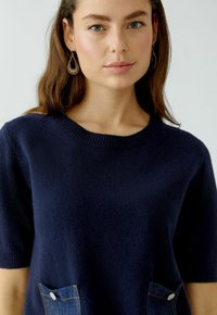 Marine gebreide sweater met korte mouwen, voorzien van denim zakken met versieringen. Gladde textuur en geribbelde halslijn voor extra detail.