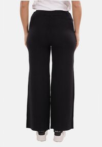 Pantaloni neri a gamba larga realizzati in un tessuto morbido con vita alta, caratterizzati da una superficie liscia e un design minimale. Indossati con sneaker bianche.
