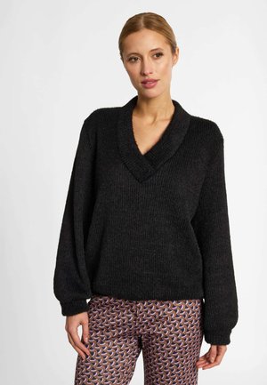 Femme aux cheveux châtain clair porte un pull noir à col en V et un pantalon à motifs avec des formes courbes marron, roses et violettes.