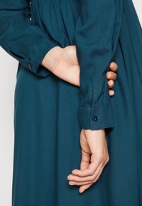 Tyrkysové šaty mají dlouhé rukávy s knoflíkovými manžetami. Látka má hladkou texturu. Jedna ruka spočívá za druhou v pase.