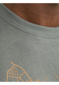 Camiseta de algodón gris con un cuello acanalado, que presenta un diseño de mapa naranja impreso en el frente. Textura suave, ajuste casual.