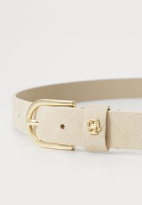 Curea din suede bej, cu o cataramă metalică aurie, având o textură netedă și mai multe găuri de ajustare. Detaliu mic cu logo embosat inclus.