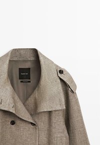 Beige dobbeltradet trenchcoat med knappet skulderrem og bred krave, mærket Massimo Dutti, vist på hvid baggrund.