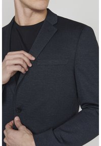 Czarny szary żakiet wykonany z teksturowanej tkaniny. Posiada zapięcie na jeden guzik, kieszeń na piersi oraz klapy o wykroju notcha. Zbliżenie na przód.