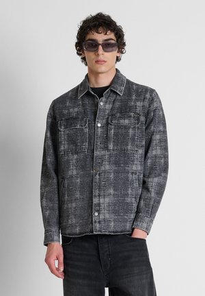 Junger Mann trägt dunkelgraue, strukturierte Jeansjacke, schwarze Hose, Sonnenbrille und steht vor einem schlichten hellgrauen Hintergrund mit einer Hand in der Tasche.