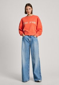 Felpa corallo con testo 'PEPE .FANS' in crema, vestibilità oversize. Abbinata a jeans a gamba larga di colore blu chiaro, che mostrano una silhouette rilassata.