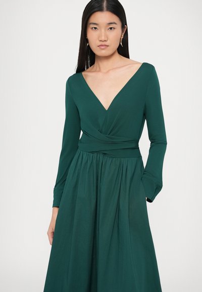 Πράσινο φόρεμα με μακριά μανίκια, βαθύ V ντεκολτέ, λεπτομέρεια στριφτού μπούστου και ρέον φούστα. Κατασκευασμένο από απαλό ύφασμα με τσέπες.