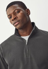 Pile grigio scuro con cerniera a un quarto e collo alto, caratterizzato da una texture liscia e una maglietta bianca visibile sotto.