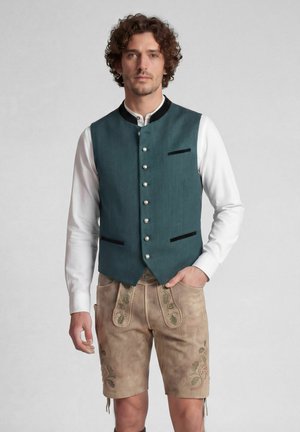 Mann mit lockigem Haar, der traditionelle bayerische Tracht trägt, darunter eine türkise Weste, weißes Hemd und bestickte Lederhosen.