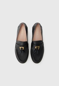 Mocassins en cuir noir avec un accent en métal doré et un détail de pampille. Texture lisse, forme arrondie et design minimaliste.