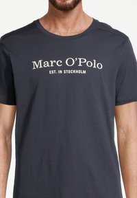 Navyblå bomulds t-shirt med korte ærmer og rund hals. Foran står "Marc O'Polo" og "EST. I STOCKHOLM" i hvid tekst.