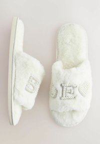 White fluffy slide sandals feature a soft texture, adorned with beaded letters 'B' and 'E' and a heart. The sole is flat and cushioned.