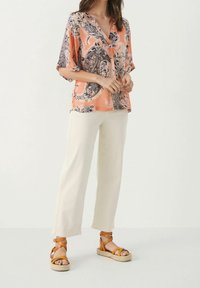Femme portant un blouse ample en motifs paisley pêche, un pantalon large écru et des sandales à plateforme couleur camel avec des brides cheville, se tenant devant un fond blanc.