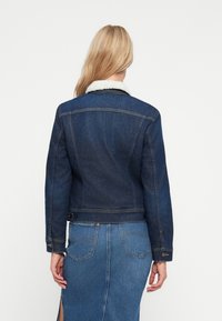 Giacca di jeans blu scuro con un colletto in fleece bianco. Caratterizzata da un design strutturato, cuciture visibili e una chiusura a bottoni ai polsini.