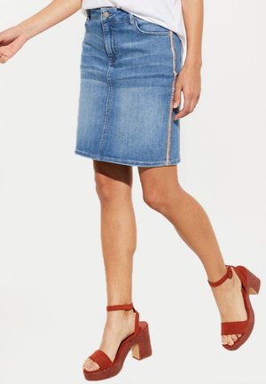 Ołówkowa spódnica dżinsowa o długości do połowy łydki, jasnoniebieskiego koloru, z widocznym przednim zamkiem błyskawicznym i bocznymi przeszyciami. Noszona z brązowymi sandałami na platformie.