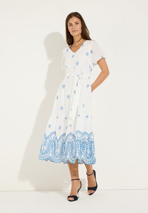 Frau trägt weißes Midikleid mit blauen Blumenmustern, kurzen Ärmeln, Taillengürtel und schwarzen Sandalen mit hohen Absätzen, steht vor einer weißen Wand.