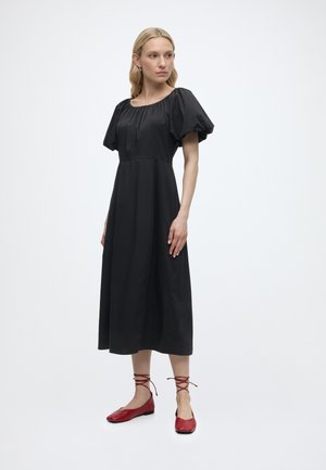 Naine, kes kannab musta midi-kleitmütsiga pluusiga ning punast paeltega baleriinisussid, seisab valge ühevärvilise tausta ees.