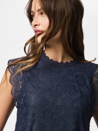 Hosszú barna hajú nő, aki tengerészkék csipkés ujjatlan felsőt visel, és balra néz egy egyszínű fehér háttér előtt.