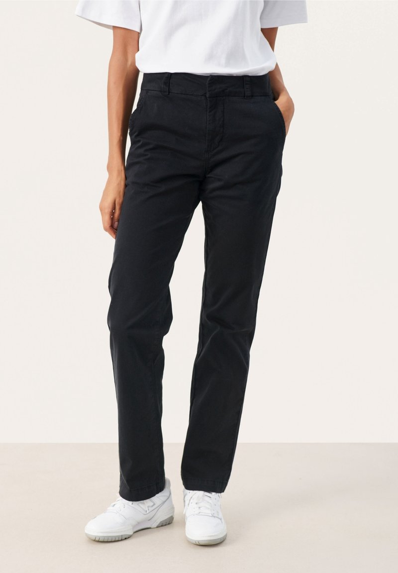 Pantalon en coton noir à coupe droite, avec poches avant et taille mi-haute, associé à des chaussures de sport blanches.