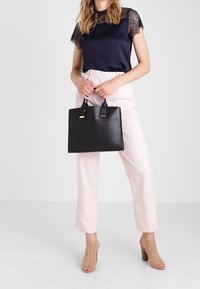 Kvinna i marinblå topp med spetsärmar och ljusrosa byxor som håller en svart handväska, bärande beige öppetåade klackskor, stående mot en enfärgad bakgrund.