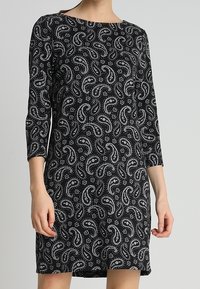 Abito nero con un motivo paisley bianco. Maniche lunghe, scollatura tonda, lunghezza sopra il ginocchio, realizzato in un tessuto morbido e elasticizzato.