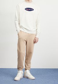 Vit sweatshirt med en rundad navy- och röd "NIKE"-logotyp, ihop med beiga sweatpants och vita hög-top sneakers. Släta tyger och avslappnad passform.