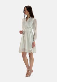 Vestido blanco de manga larga con cuello y cintura ajustable. Tejido transparente con una capa interior opaca, combinado con sandalias de tacón alto con tiras.