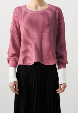 Personne portant un pull en maille côtelée rose ample par-dessus une chemise blanche à manches longues et une jupe plissée noire, debout devant un fond uni.