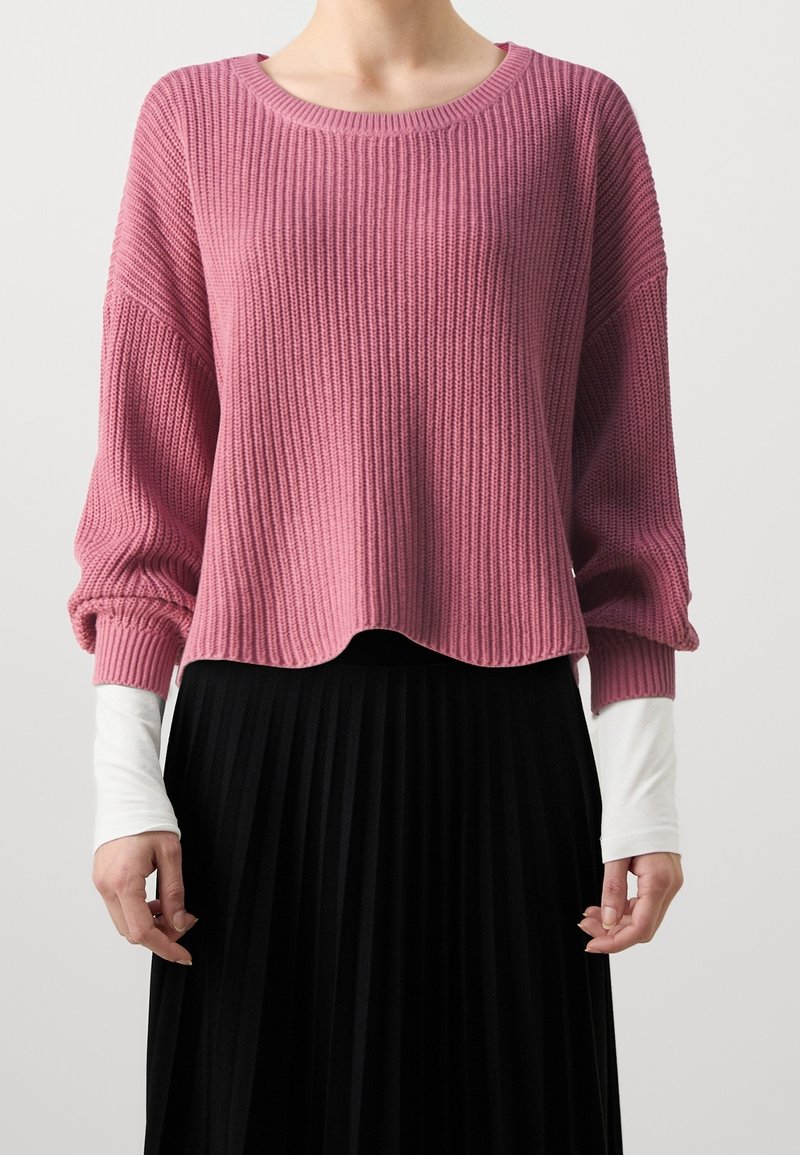 Personne portant un pull en maille côtelée rose ample par-dessus une chemise blanche à manches longues et une jupe plissée noire, debout devant un fond uni.