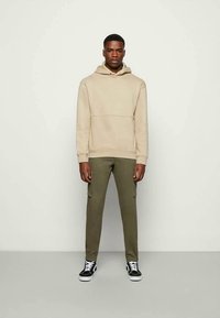 Uomo in piedi contro uno sfondo neutro, che indossa un felpa beige, pantaloni cargo verde oliva e sneakers nere con lacci bianchi.