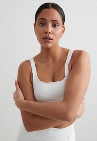 Hvit ribbet tank topp med tykke stropper, med firkantet utringning. Modellen krysser armene mot en ensfarget bakgrunn.