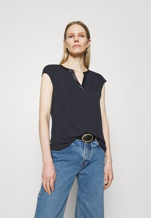Schwarzes kurzärmliges Oberteil mit V-Ausschnitt und glänzender Paspel, kombiniert mit hochgeschnittenen blauen Jeans, die einen schwarzen Gürtel mit einer runden goldenen Schnalle tragen.