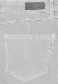 Poche arrière d’un pantalon en velours côtelé gris clair avec des surpiqûres visibles et une petite étiquette en cuir rectangulaire de marque "OPUS" au-dessus de la poche.